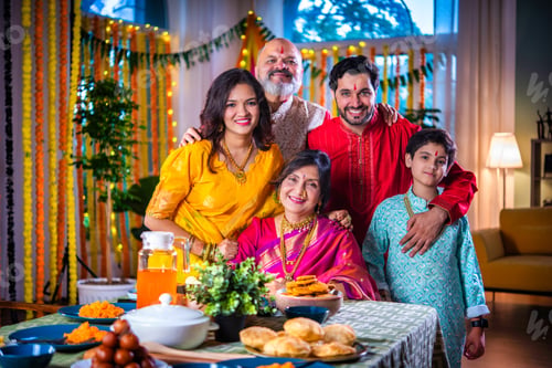 Preview: Portrait of happy Indian family in traditional outfit celebrating Diwali at home eating food