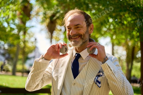 Preview: Smiling man holding headphones in a sunny park