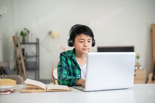 Preview: Asian kid elementary school boy wearing headphones looking at white laptop on table with book aside.