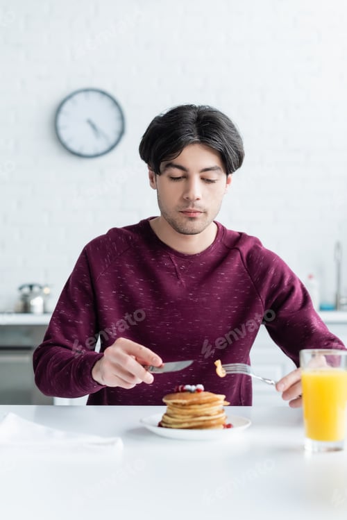 Preview: brunette man eating tasty pancakes near blurred glass of orange juice
