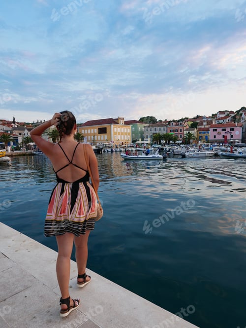 Preview: Rear view of young woman in summer dress standing in port of picturesque seaside town.