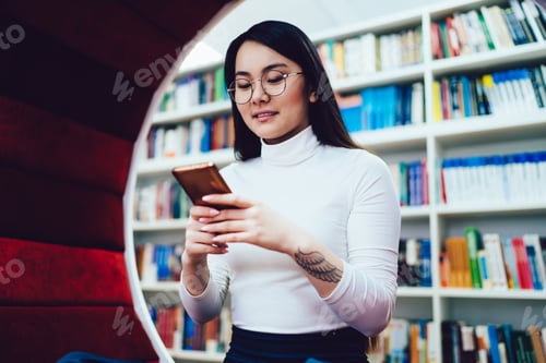 Preview: Smiling Asian woman with tattoo on hand browsing smartphone in library