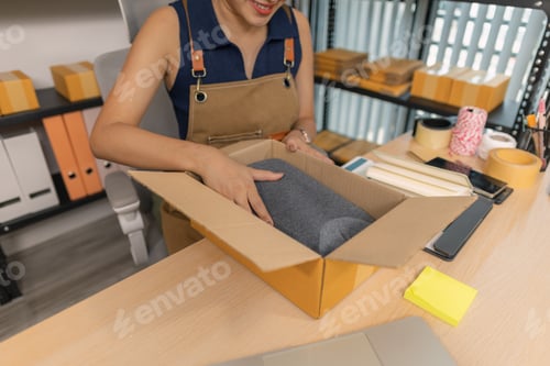 Preview: Smiling young female entrepreneur packing clothes into a parcel box