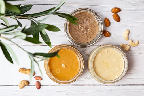 Preview: Jars of Almond, Cashew and Peanut Butter on a White Wooden Table.