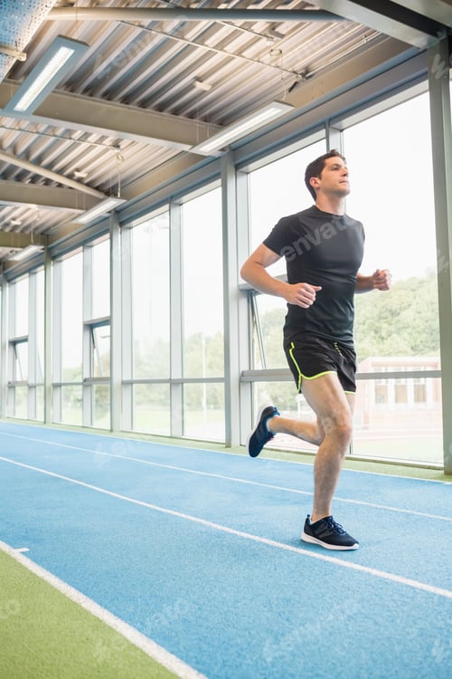 Preview: Male athlete running on blue synthetic track lanes by glass panels at athletic center, copy space