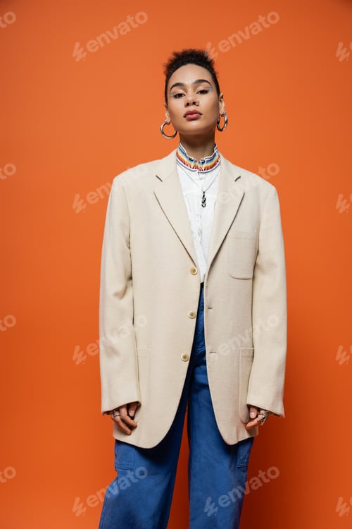 Preview: trendy young fashion model in beige blazer and jeans with hoop earrings posing on orange backdrop