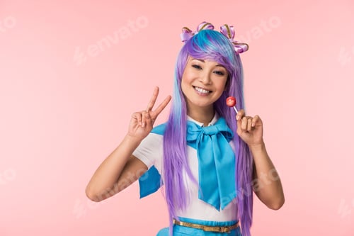 Preview: Happy asian anime girl holding lollipop and showing peace sign isolated on pink