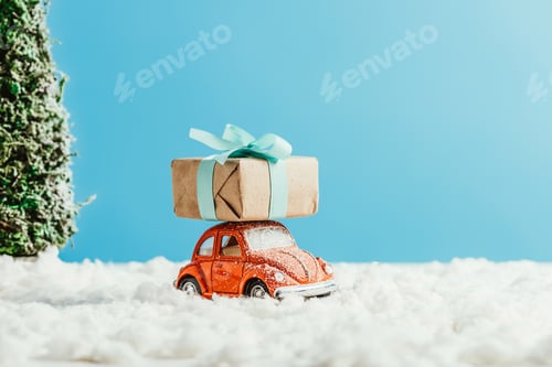 Preview: close-up shot of toy red car with christmas gift box riding by snow made of cotton on blue