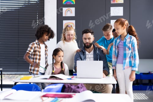 Preview: Diverse elementary schoolchildren around male teacher using laptop at desk in classroom, copy space