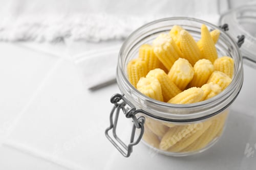 Preview: Tasty fresh yellow baby corns in glass jar on white tiled table, closeup. Space for text