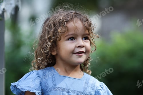 Preview: A cute little curly girl in a blue dress stands in the garden on a summer day, looking away,portrait