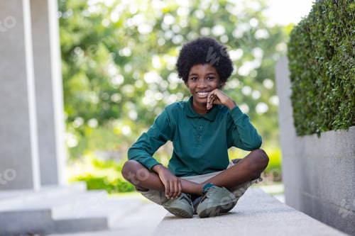 Preview: A black curly haired African boy sits smiling with his hands on his chin and looks at the camera