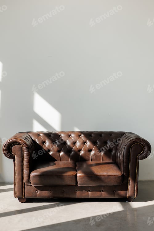 Preview: Vintage quilted classic brown sofa against white wall with sunlights from window on concrete floor