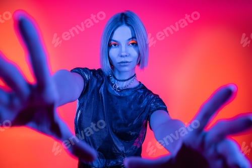 Preview: stylish woman with outstretched hands looking at camera in blue neon light on coral and pink