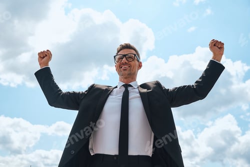 Preview: Happy mature man in full suit keeping arms raised while standing in front of blue sky outdoors