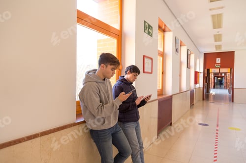 Preview: two university student friends leaning against the wall using mobile phones
