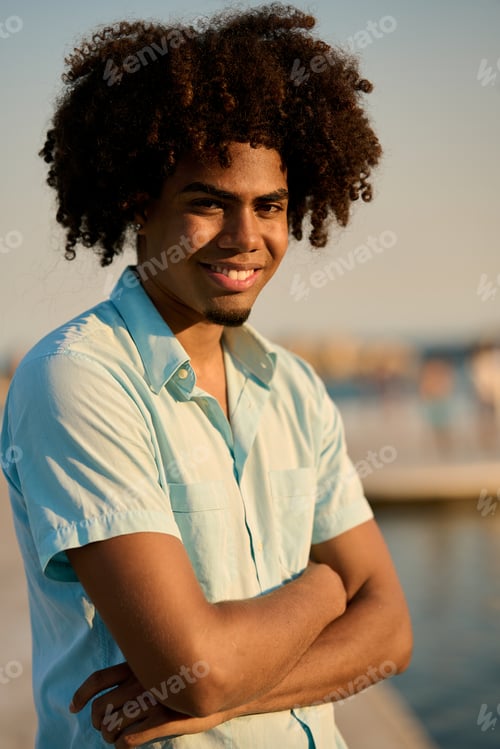 Preview: Young man smiling with folded arms by the sea at sunset