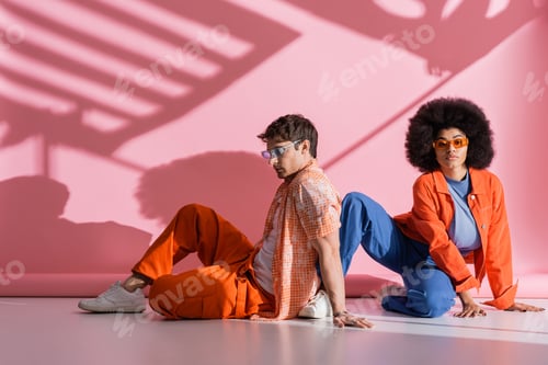 Preview: Stylish interracial couple in sunglasses sitting on pink background with shadow