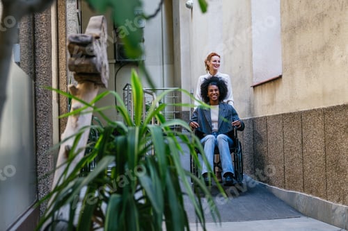 Preview: Two young businesswomen entering coworking space, one using wheelchair ramp