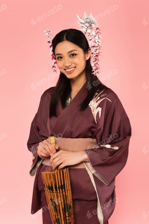 Preview: Smiling japanese woman with umbrella looking at camera isolated on pink