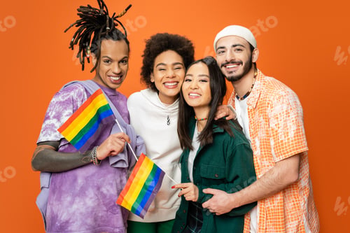 Preview: multicultural friends in stylish clothes holding lgbt flags and smiling at camera isolated on orange
