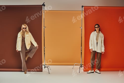 Preview: autumn fashion, stylish multicultural models in sunglasses and outerwear posing on colorful backdrop