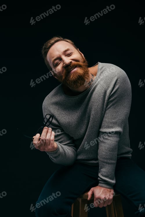 Preview: Smiling bearded man in jumper holding eyeglasses while sitting on chair isolated on black