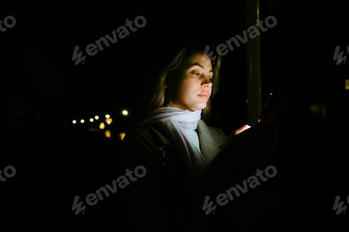 Preview: Young caucasian woman using mobile phone outdoor in the evening, walking on city streets.