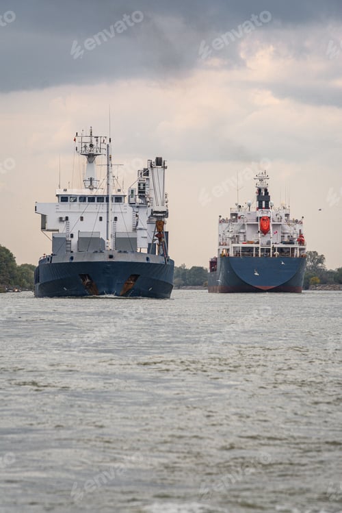 Preview: Cargo ships navigating a river under a cloudy sky.