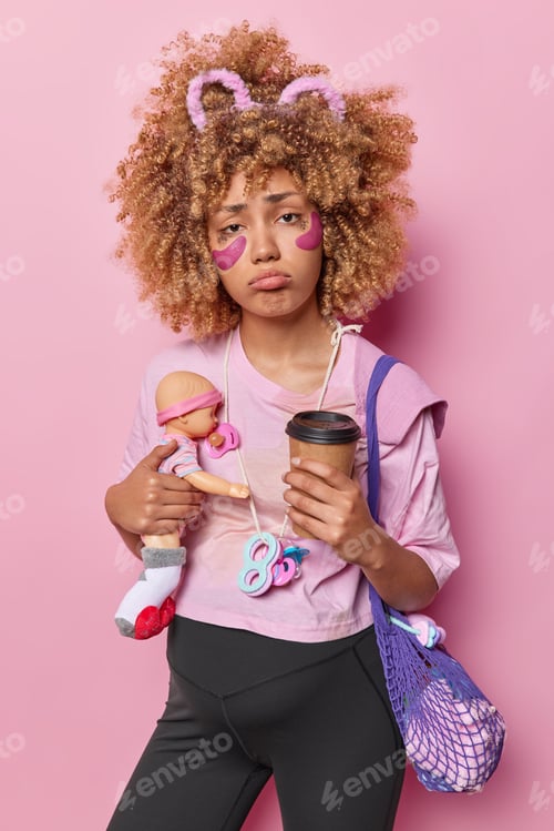 Preview: Image of frustrated young curly haired woman has unhappy sad expression holds doll and paper cup of