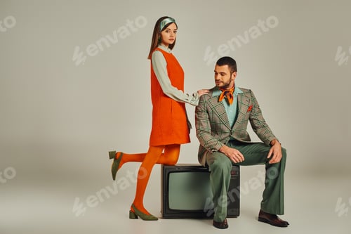 Preview: woman in orange dress touching shoulder of man sitting on vintage tv-set on grey, retro style