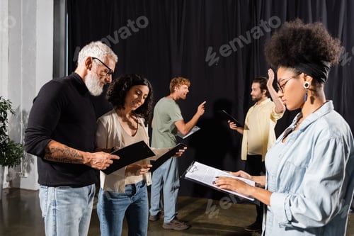 Preview: african american woman reading screenplay near bearded art director and troupe rehearsing in theater