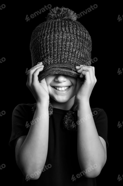 Preview: Vertical grayscale shot of a happy little boy covering his eyes with a fluffy winter hat and smiling