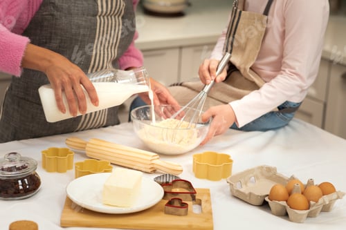 Preview: European adult lady, little girl in apron make cookie dough together, poure milk, flour and eggs