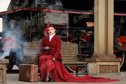 Preview: A girl in a burgundy suit and a hat with a veil is sitting on a suitcase near an old locomotive