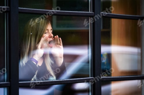 Preview: portrait of middle aged blonde woman staying behind window wth reflections, breathing on the glass