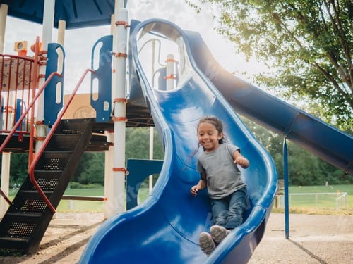 Preview: happy mixed race preschooler having fun outdoors on a blue slide at the park