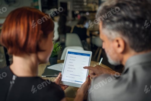 Preview: Rear view of man and woman having business meeting in a cafe, using tablet.