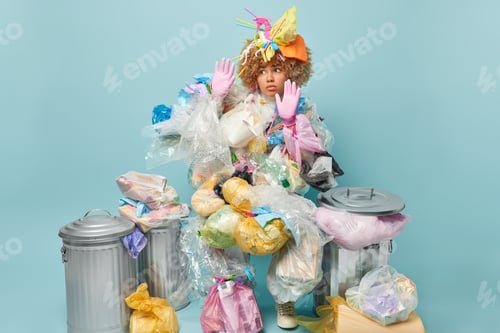 Preview: Serious curly haired woman covered with plastic garbage keeps palms raised up tries to protect