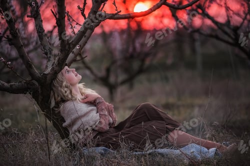 Preview: Young beautiful curly blonde woman sits in blooming peach sunset gardens