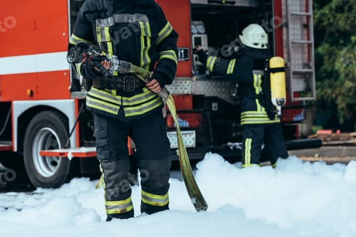 Preview: cropped shot of firefighter in water hose in hands standing in foam on street