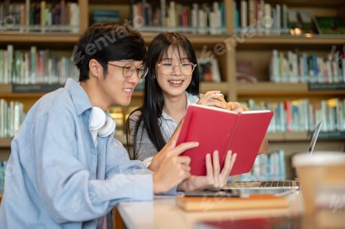Preview: Glasses asian man student holding book reading with a woman friend while sitting at library's table.