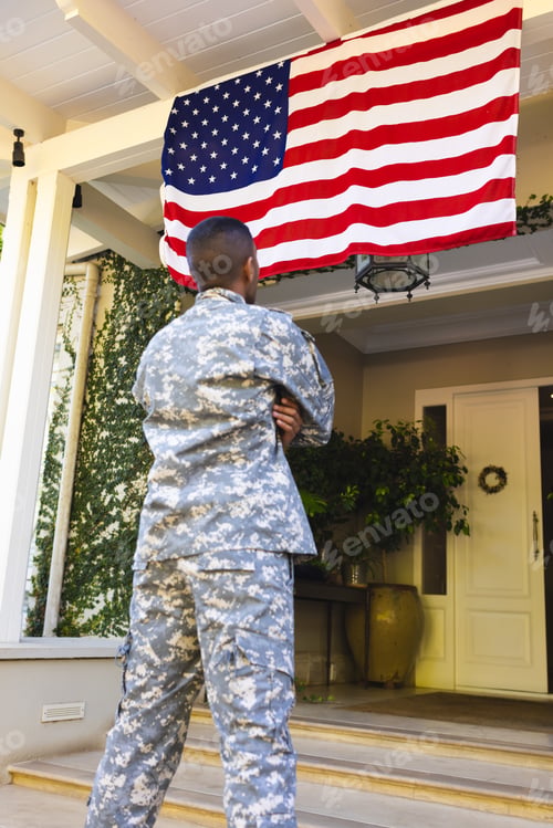 Preview: Biracial male american soldier wearing military uniform looking at flag of usa outside the house