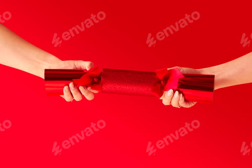 Preview: Close Up Studio Shot Of Female Hands Pulling Christmas Cracker Against Red Background