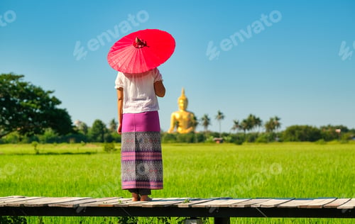 Preview: Woman with traditional skirt and red umbrella stand on wood bridge in front of green rice field