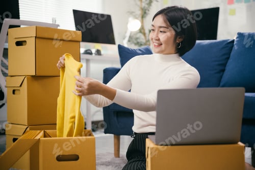 Preview: Asian female entrepreneur preparing clothes for shipping using laptop at home office