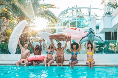 Preview: Happy group of friends enjoying swimming pool party at the resort holding inflatables.