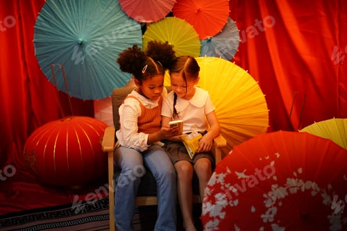 Preview: Two little girls looking at smartphones with red Chinese umbrella background