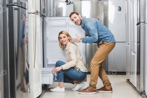 Preview: smiling boyfriend showing like and girlfriend sitting near new fridge in home appliance store