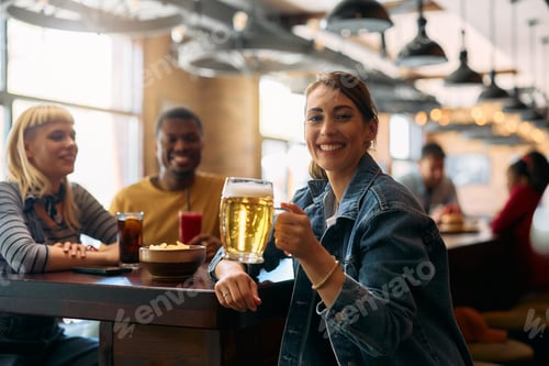 Preview: Happy woman drinking beer with friends in a pub and looking at camera.